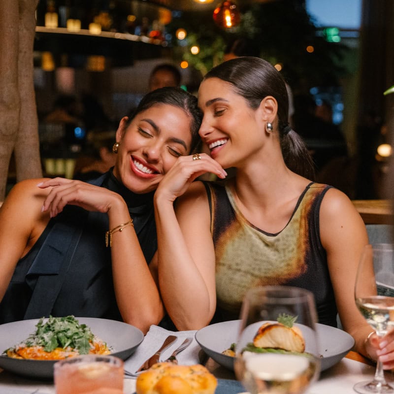 One female leaning on another female, both are smiling in a restaurant setting
