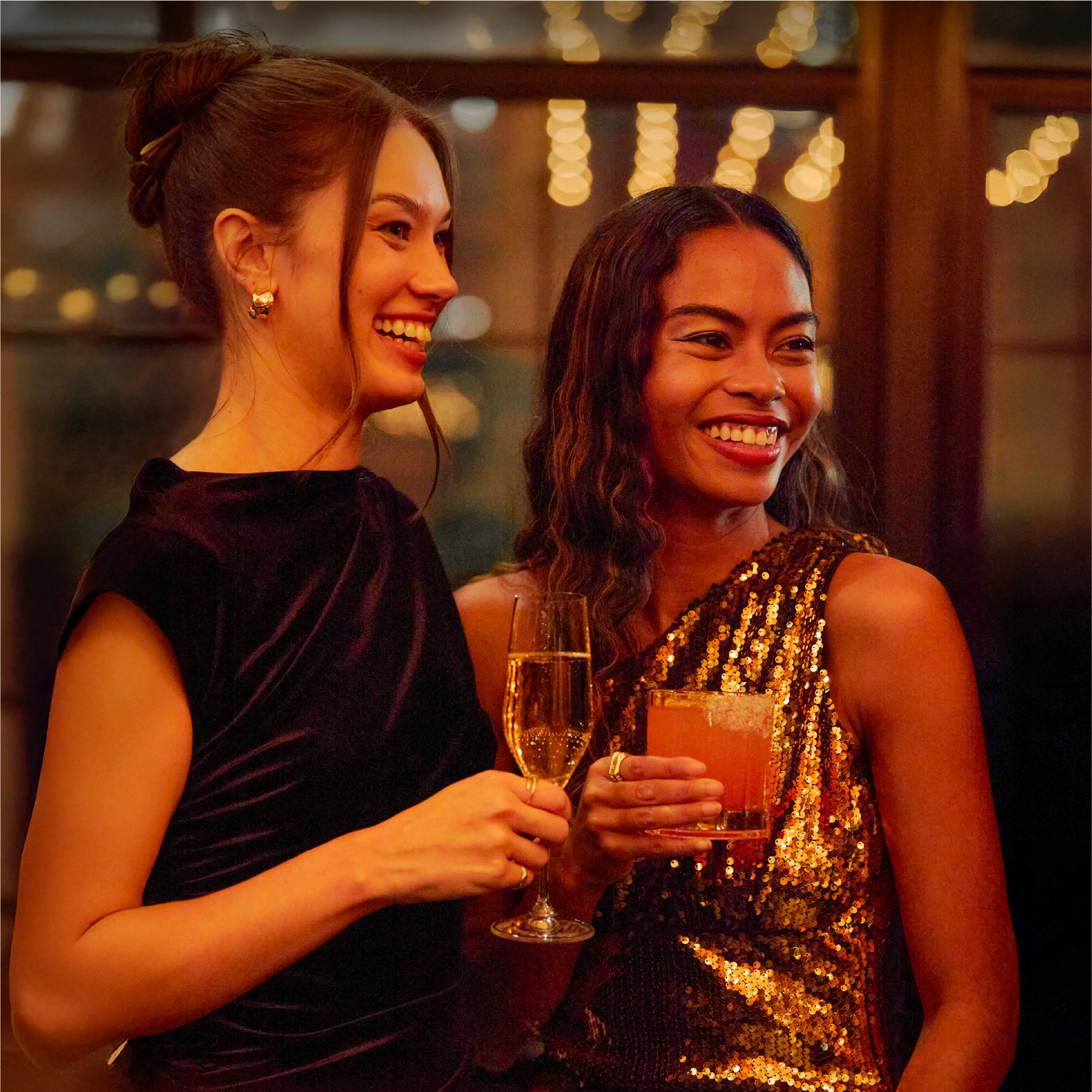 Two stylish women smiling and holding cocktails at a holiday party